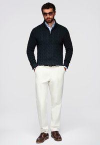Homme portant des lunettes de soleil, un pull noir en tricot torsadé, une chemise bleu clair à col, un pantalon blanc et des chaussures en cuir marron, se tenant les mains dans les poches.