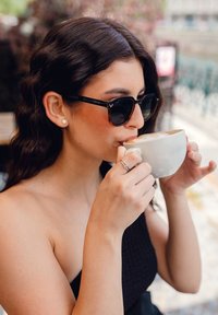 Černé a želvovinové sluneční brýle čtvercového tvaru. Bílý keramický hrnek držený oběma rukama, naplněný krémovým nápojem.