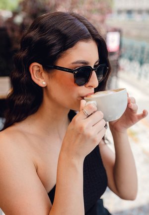 Černé a želvovinové sluneční brýle čtvercového tvaru. Bílý keramický hrnek držený oběma rukama, naplněný krémovým nápojem.