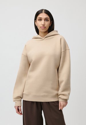 Beige oversized hoodie made of soft fabric; features a drawstring hood, ribbed cuffs, and waistband; smooth texture. Paired with dark trousers.