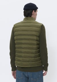 Maslinasta pufasta prsluk s klesanim dizajnom, rebrastim pletenim detaljima i ravnim rubom, složen preko teksturirane tamnozelene veste.