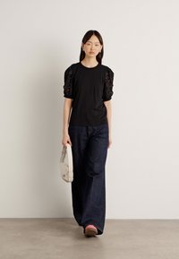 Μαύρο t-shirt με μανίκια από δαντέλα, συνδυασμένο με σκούρες τζιν παντελόνες σε φαρδιά γραμμή και ροζ υπόδηση. Το μοντέλο κρατάει μια μπεζ τσάντα ώμου.