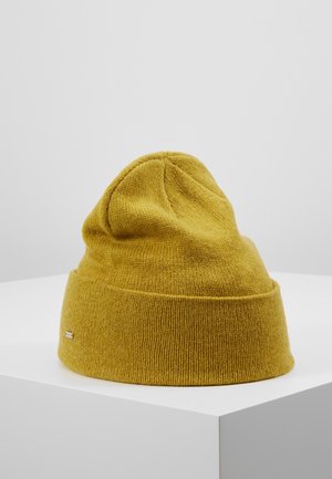 Mosterdgele gebreide beanie met een opgerolde boord, tentoongesteld op een wit voetstuk tegen een effen lichtgrijze achtergrond.