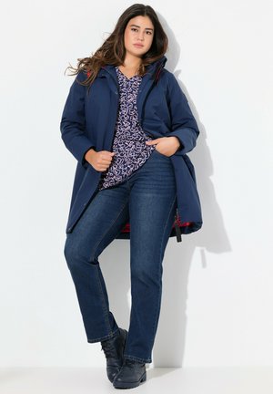Navy waterproof coat with red accents, worn over a patterned top and paired with blue jeans and black boots, featuring a relaxed fit.