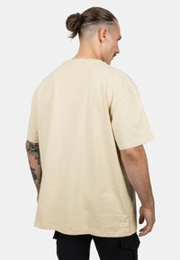 Beige cotton t-shirt with short sleeves and a relaxed fit, showing a smooth texture and minimal seams, viewed from the back.