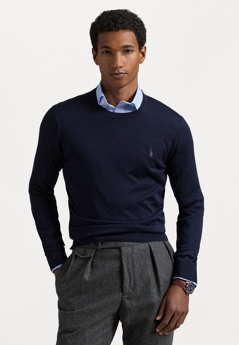 Pull à col rond bleu marine avec un tricot texturé, associé à une chemise à col bleu clair. Pantalons plissés gris et une montre avec un cadran bleu.