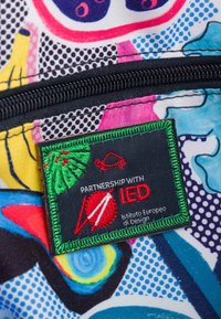 Tela colorida con patrones abstractos en azul, rosa y amarillo; incluye una etiqueta bordada en verde con la inscripción "Asociación con IED."