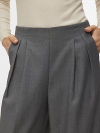 Femme portant un pantalon plissé gris taille haute, mains dans les poches, et un haut beige clair à manches longues rentré.