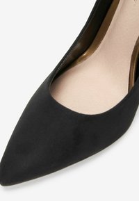 Czarne zamszowe buty na wysokim obcasie z szpiczastym czubkiem i gładką, beżową wkładką. Wnętrze ma metaliczny akcent wzdłuż krawędzi.