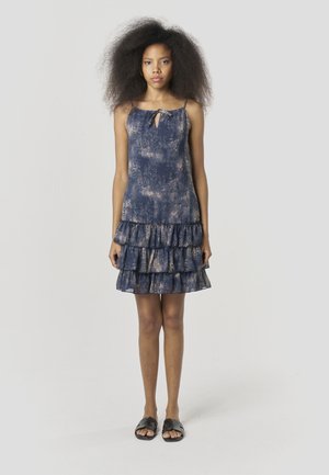 Vestido azul marino sin mangas con dobladillo con volantes y detalle de cuello con lazo. La tela tiene un acabado texturizado y jaspeado. Se lleva con sandalias planas negras.