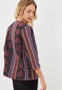 Mujer vestida con una chaqueta negra con rayas verticales de colores y patrones florales rojos, mirando hacia abajo sobre su hombro contra un fondo blanco.