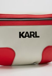 Bolso de cuero blanco y rojo con cremallera, que presenta un logo negro de "KARL" y acentos rojos en los laterales. Textura lisa con bordes redondeados.