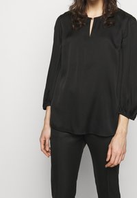 Blouse noire à manches longues et bouffantes, détail au niveau du col en forme de trou de serrure, et texture lisse et brillante. Associée à un pantalon noir assorti.
