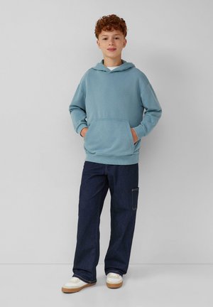Ragazzo con capelli ricci rossi in piedi con le mani nelle tasche, indossando una felpa leggera blu, jeans blu scuro e sneakers bianche su uno sfondo semplice.