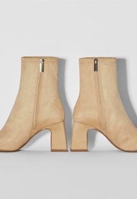 Stivaletti ankle in suede beige con tacco quadrato, caratterizzati da una cerniera laterale e dettagli cuciti. Texture liscia e design minimalista.