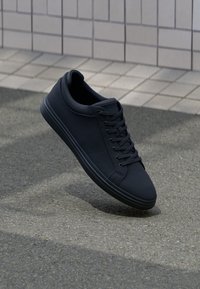 Black textured sneakers with a rounded toe, flat laces, and a minimalist design. The sole is smooth and slightly elevated.