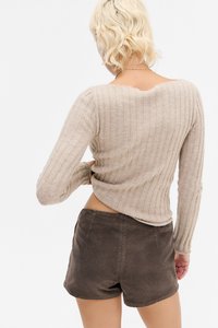 Blonde persona ar īsiem viļņainiem matiem, tērpusies bēšā ribotā džemperī un tumši brūnos kordūru šortos, stāv ar muguru pret skatītāju, pielāgojot džempera maliņu.
