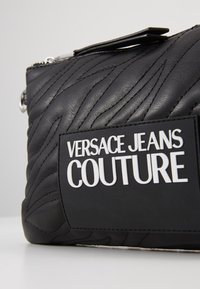 Svart läderväska med quiltad textur och upphöjda detaljer, en djärv vit logopatch och silverfärgade detaljer. Dragkedjestängning.