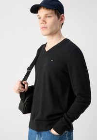 Pulover negru cu decolteu în V, cu mâneci lungi, textură moale, logo mic pe piept, asociat cu blugi albaștri și o geantă de umăr neagră.