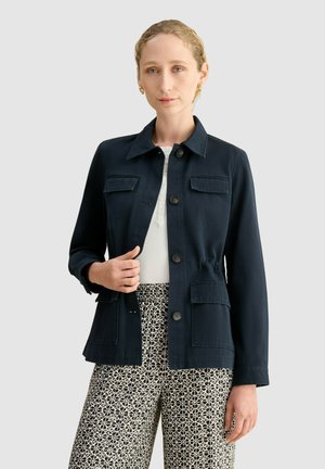 Mujer con chaqueta negra abotonada sobre un top blanco y pantalones anchos con estampado, de pie frente a un fondo liso y claro.