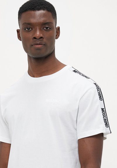 Vit bomull t-shirt med rund halsringning, korta ärmar och svart "MOSCHINO"-text längs axelsömmen. Texturerad logotyp på bröstet.