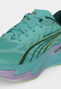Gros plan d'une chaussure de course turquoise avec des lacets turquoise, une bande noire et jaune, et une semelle violette, avec le texte "MY SPORT IS MY RO".