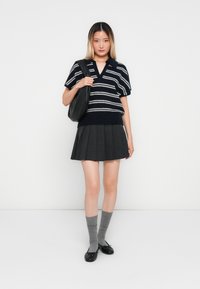 Femme portant un pull à rayures noir et blanc à manches courtes, une jupe plissée noire, des chaussettes grises, des ballerines noires et portant un sac à bandoulière noir.
