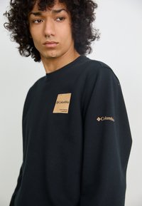 Sudadera negra de algodón, con un parche texturizado de color marrón claro en el pecho y la marca Columbia en marrón claro en la manga.