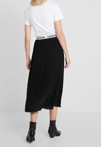 Gonna maxi nera con logo Calvin Klein Jeans in vita, texture morbida, spacco laterale, abbinata a una t-shirt bianca aderente e stivaletti neri alla caviglia.