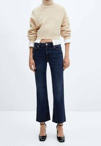 Jeans évasés en denim bleu foncé à taille haute, avec des poches avant et un détail de couture subtil, associés à un pull court beige.