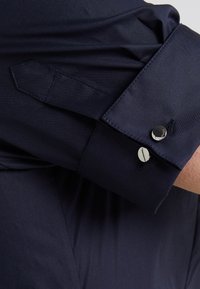 Polsino di una camicia blu scuro con due bottoni argentati, uno incisi con "Hugo," e un dettaglio sulla manica su un braccio piegato.