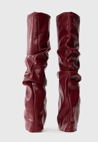 Botas altas y holgadas hasta la rodilla en cuero rojo oscuro liso con un diseño fruncido, de pie sobre un fondo blanco.