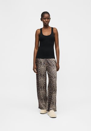 Femeie purtând un maiou negru, pantaloni largi cu imprimeu de leopard și pantofi bej slip-on, stând pe un fundal alb simplu.