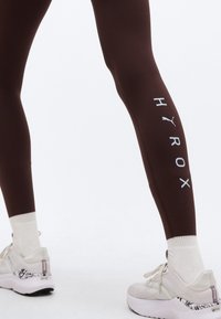 Πόδια με σκουρό καφέ κολάν που έχουν λευκό λογότυπο «HYROX» και Puma, συνδυασμένο με λευκές κάλτσες και λευκά παπούτσια Puma με μοτίβο στη φτέρνα.