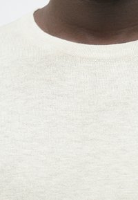 Κοντινό πλάνο ενός σκούρου δέρματος ατόμου που φοράει ένα κρεμ τύπου πουλόβερ με στρογγυλή λαιμόκοψη.