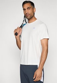 Biela športová tričko s krátkymi rukávmi, na hrudi s malou nášivkou. Model drží raketu a má na sebe tmavosivé nohavice.