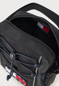 Borsa di stoffa nera con zip, caratterizzata da dettagli blu, un cordino e una patch con logo. Interno visibile, che mostra un ampio scomparto.