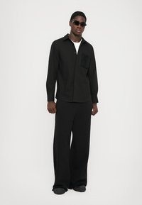 Chemise noire avec une poche texturée, manches longues et fermeture à boutons. Associée à un pantalon noir à jambes larges et des chaussures noires.