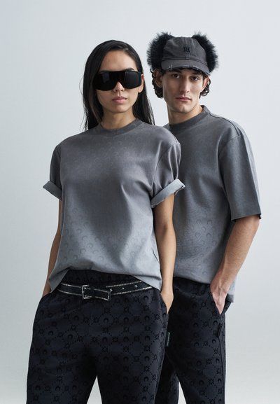 Graue Kurzarm-T-Shirts mit aufgerollten Ärmeln und einem dezenten Muster. Dunkle Hosen mit einem ähnlichen Muster und einem mit Nieten versehenen Gürtel. Accessoires sind große schwarze Sonnenbrillen und eine graue Mütze mit Fellbesatz.