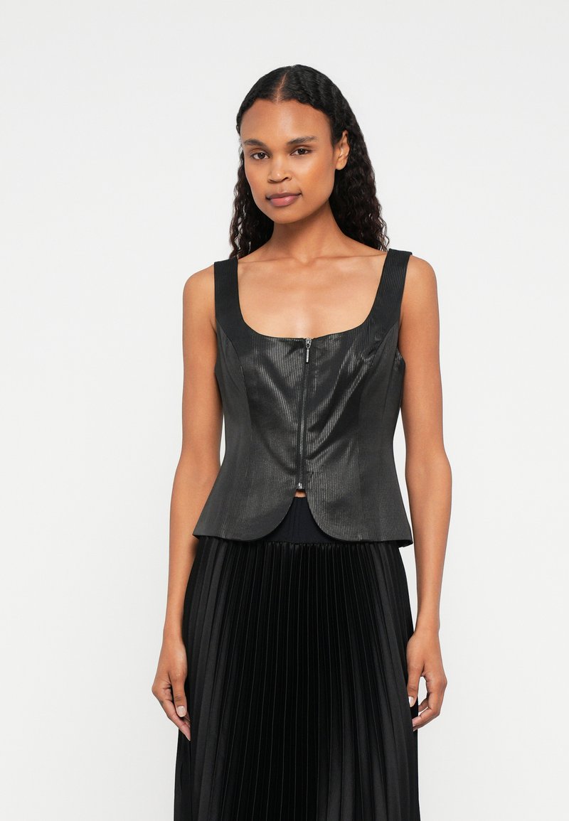 Femme avec de longs cheveux bouclés portant un haut sans manches zippé noir et une jupe plissée noire, se tenant devant un fond blanc.