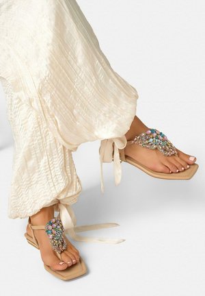 Pies con sandalias beige de punta cuadrada con adornos de joyas multicolores y pantalones crema plisados atados en los tobillos.