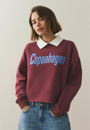 Junge Frau mit kurzen braunen Haaren trägt einen kastanienbraunen Pullover mit der Aufschrift "Kopenhagen" in Blau, dazu ein weißes Hemd mit Kragen und eine blaue Jeans.