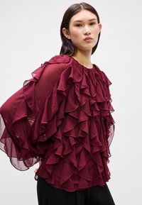 Maroon blouse made of lightweight fabric, featuring multiple ruffles along the front and sleeves, round neckline, and loose fit.