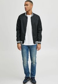 Giacca bomber nera con colletto e polsini a righe bianche e nere, abbinata a una maglietta bianca e jeans blu. Calzature sportive.