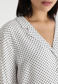 Vit blus med ett svart stjärnmönster, med krage och V-urringning. Tyget har en slät, lätt struktur.