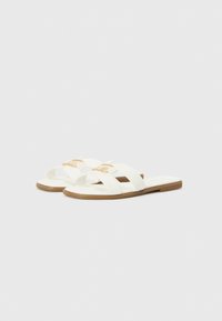 White sandals with three wide straps, featuring a gold buckle accent and a flat brown sole. Smooth texture and minimalistic design.