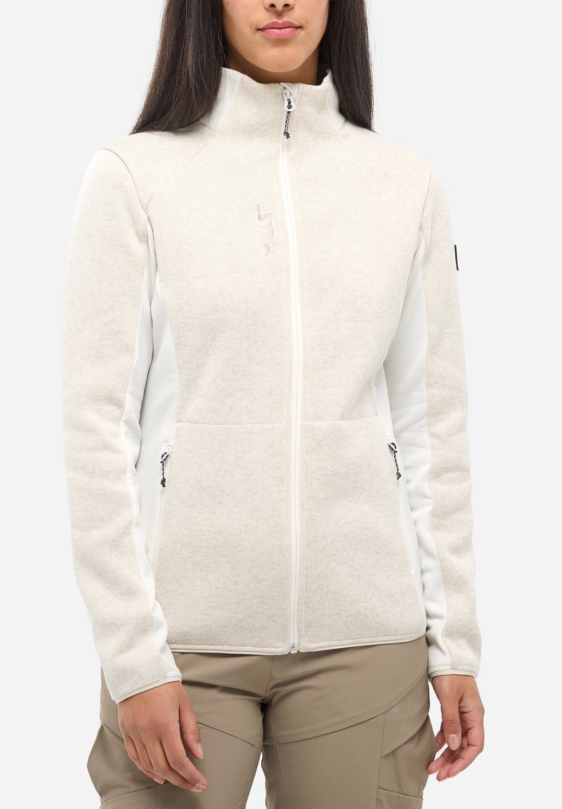 Frau, die eine beigefarbene Fleecejacke mit Reißverschluss und weißen Seitenpanelen sowie Reißverschlusstaschen trägt, kombiniert mit hellbraunen Hosen.