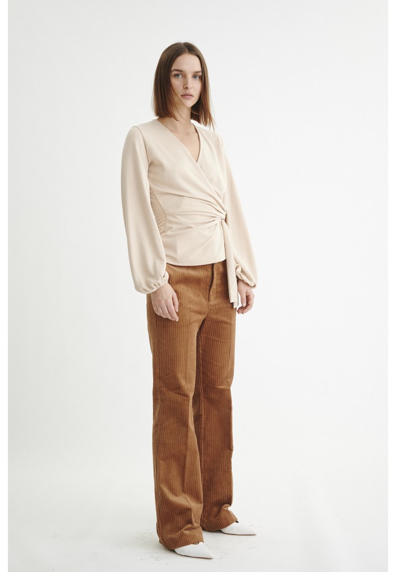 Blusa bege de manga longa com detalhe plissado na cintura, combinada com calças flare de cabedal castanho. Sapatos brancos de bico fino.