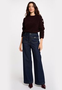 Maglione testurizzato bordeaux abbinato a jeans in denim scuro a gamba larga con tasche con doppia zip e cuciture dorate. Indossato con tacchi a punta bordeaux.