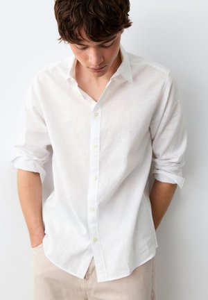 Jeune homme aux cheveux courts et bruns portant une chemise blanche boutonnée avec les manches retroussées et un pantalon beige clair, regardant vers le bas les mains dans les poches.